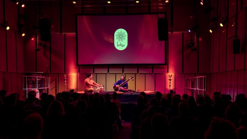 1/11/23 - Indika Festival | Wide-angle shot of the crowd watching Shahbaz Hussain and Shakir Khan performing on tabla and sitar with the Indika logo projected onto a projector screen in the Music Room in the Liverpool Philharmonic concert hall.